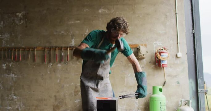 Caucasian male blacksmith wearing safety glasses, hammering hot metal tool on anvil in workshop