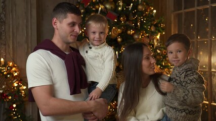 Portrait of a smiling young family with young children near the Christmas tree waving a greeting to the camera. Happy parents with young children cheerfully celebrate New Year's holidays