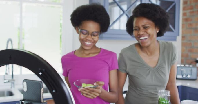 Happy african american mother and daughter preparing healthy drink, making video using smartphone