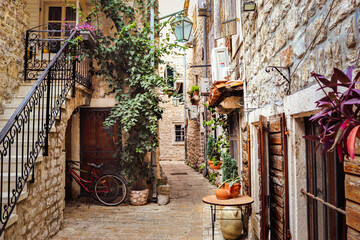 Cozy narrow Roman street in the old town of Budva. Flowers, plants, old bicycle, street lamp, wooden shutters. Montenegro, Balkans. Summer.