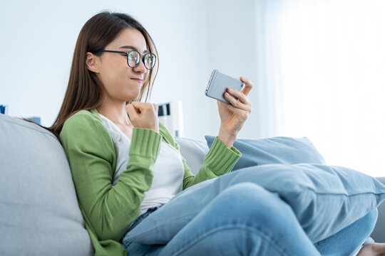 Asian Woman Wear Eyeglasses Play Mobile Game On Smartphone At Home. 