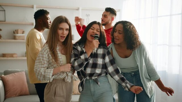 Karaoke party at home. Group of happy multiethnic women singing together into microphone, guys dancing on background