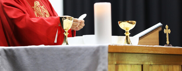 A Catholic Priest about to break the bread host while celebrating blessed Holy Communion at Mass. Wearing a red gown vestment and surrounded by chalice cross crucifix and bible - obrazy, fototapety, plakaty