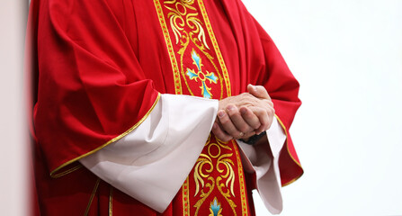 close up of a catholic priest during mass in a church wearing a bright red gold patterned vestment or robe. Standing with hands clasped, white background.