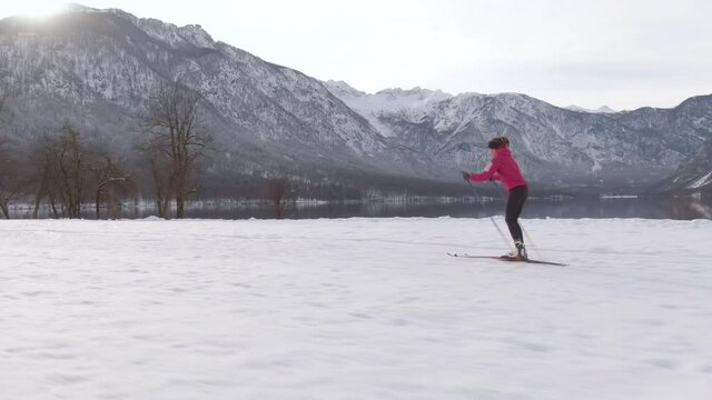 AERIAL: Drone shot of a female nordic skier training on a sunny winter day in the Slovenian mountains. Winter sun shines on the picturesque wintry lake Bohinj as a young woman trains nordic skiing.