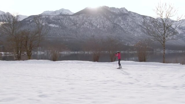 AERIAL: Scenic drone shot of a woman training nordic skiing at a picturesque ski resort in the mountains of Slovenia.. Lone Nordic skier trains near the gorgeous lake Bohinj on a sunny winter morning.