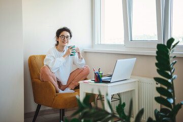 Caucasian woman sitting relaxed in armchair and drinking a cup of tea, doing her job using laptop. Modern woman lifestyle.