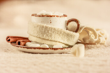 A white mug of hot cocoa with marshmallows on soft white background. A mug with knitted scarf,cinnamon sticks and marshmallows on the saucer.  Hot cocoa, coffee on a cold day.