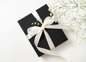 a black gift box with a white ribbon on a white background with a bouquet of  gypsophila. The concept of a holiday or gift card. Top  view and copy space