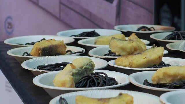 chef adds pieces of fried fish to spaghetti with squid ink on wooden plates for tasting at gastronomic event