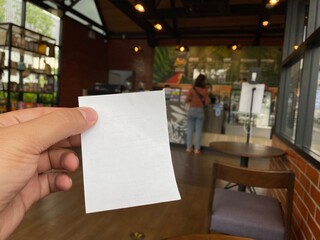 Hand holding blank white cue card with blurred cashier counter coffee shop background, Young man...