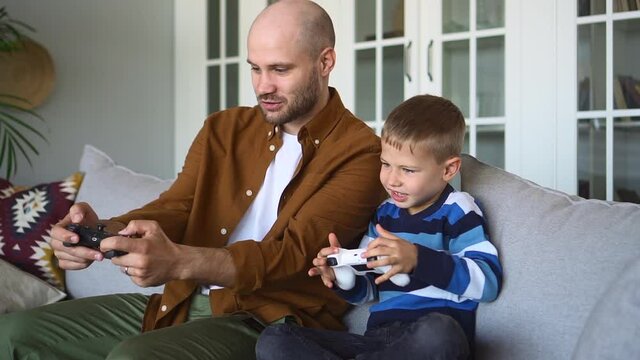 Father and toddler son have fun. Excited bearded parent shows buttons on joystick to funny child playing videogame sitting on grey sofa at home spbd