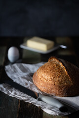 Homemade sourdough wheat bread on a dark background with an egg in a ceramic stand and butter, next to a bread knife. Dark background, a wooden table.