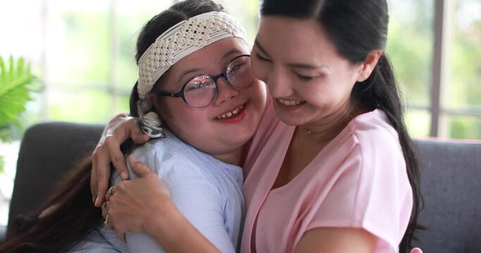 Cheerful Asian mother embracing happy daughter with down syndrome while sitting on sofa at home