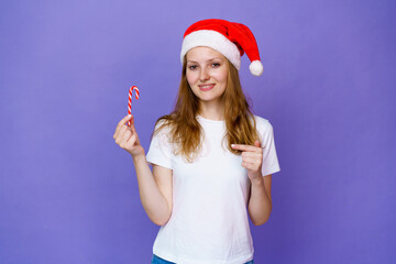 Fototapeta na wymiar Close-up of young blonde girl in santa hats and white t-shirt shows finger on lollipop in the form of a cane, on a lilac background