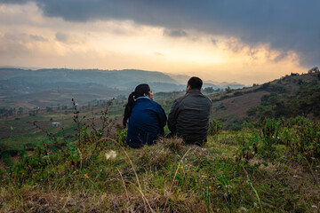Fototapeta na wymiar young couple sitting on mountain top with misty hill range background and dramatic sky at morning