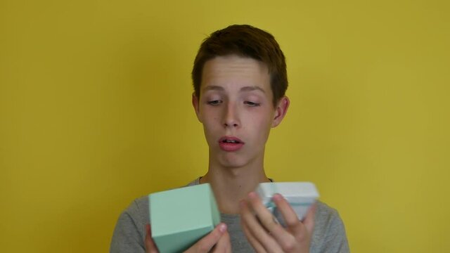 A boy 14-17 years old in a light gray T-shirt on a yellow background opens a blue gift box. The boy is disappointed with the gift. Holiday concept.