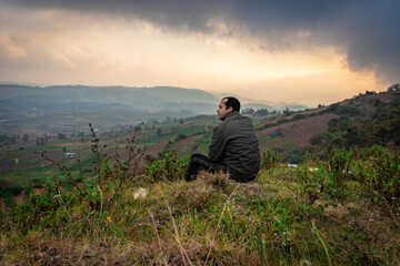 Fototapeta na wymiar young man sitting on mountain top with misty hill range background and dramatic sky at morning