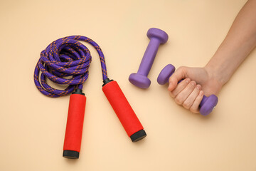 Female hand with dumbbells and skipping rope on beige background