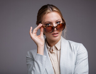 Fashion photo of a beautiful elegant young woman in a pretty soft blue suit, jacket, pants, trousers, white boots, stylish sunglasses on light gray background. Studio shot, portrait. Monochrome colors