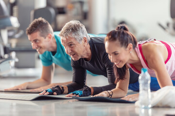 Group of people in various age and gender having fun while doing group exercise of elbow plank in the gym