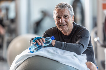 Elderly man resting on an exercise ball in the gym holds a water bottle looking at the camera