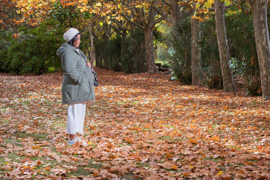 mujer embarazada con gorra blanca, abrigo gris y sosteniendo la ropa de su futuro beb&eacute; se encuentra en un parque con colores oto&ntilde;ales.