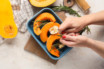 Woman putting rosemary into baking dish with fresh pumpkin pieces on light background