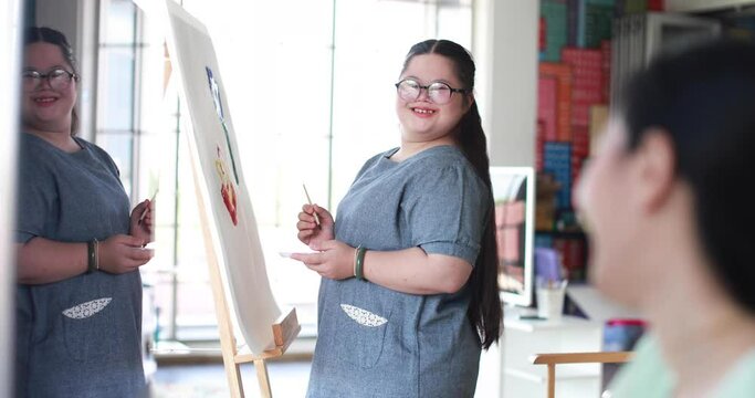 Side view of smiling Asian female teacher looking at talented girl with down syndrome painting on canvas with paintbrush during art class