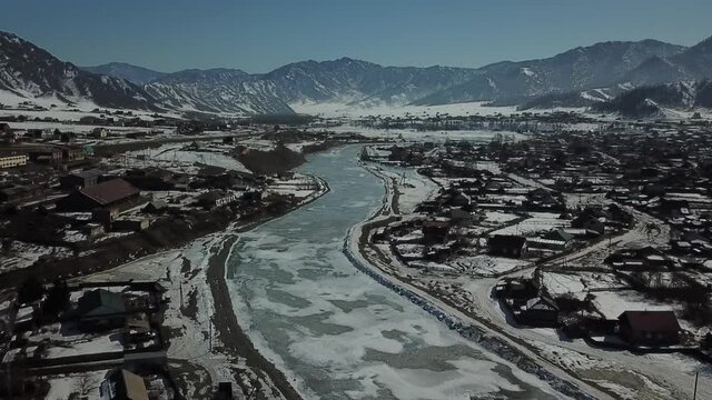 Aerial shot over iced river in forward camera movement. Village with melted snow in front of mountains