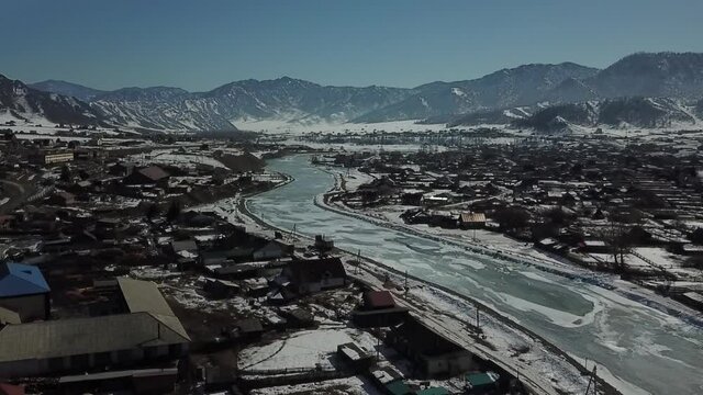 Aerial shot in forward camera movement of distant spring mountains. Village with melted snow and iced river in front of mountains