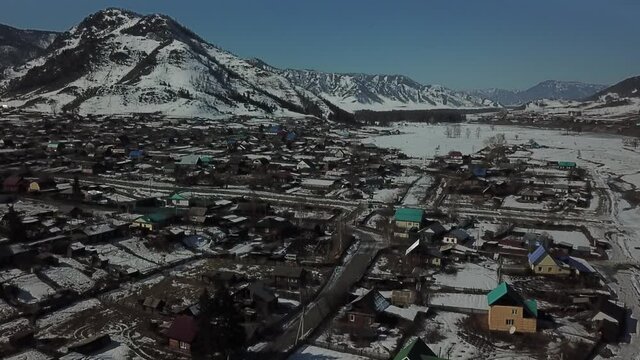 Aerial shot in forward camera movement over village to distant mountains when spring. Iced river to the right, big mountain to the left
