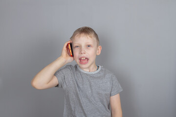 A blond Caucasian boy talks on a red smartphone and smiles on a gray background in the studio