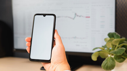 Trading, investment, business, finance and technology concept. Close-up man holding white blank screen smartphone mock-up on computer background with stock exchange graph
