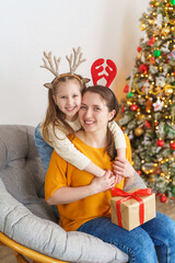 Fototapeta na wymiar loving mother and daughter dressed in reindeer horns hug at home against background Christmas tree. little girl with deer antlers on her head is happy and laughing. hugging, holding Christmas gift box
