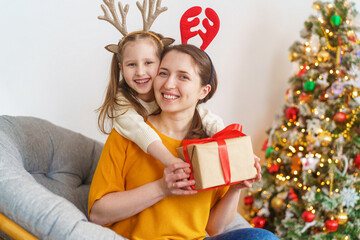 Obraz na płótnie Canvas loving mother and daughter dressed in reindeer horns hug at home against background Christmas tree. little girl with deer antlers on her head is happy and laughing. hugging, holding Christmas gift box