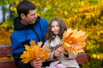 Father and daughter portrait in an autumn park. Happy people pose against the background of beautiful yellow trees. They sit on a bench, hug and are happy together.