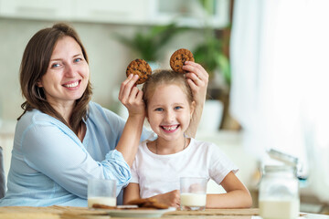 Obraz na płótnie Canvas happy loving family, mom and daughter, playing sitting at table and having breakfast at home in morning. woman and girl eat oatmeal cookies and drink cow's milk, and have nice time together in kitchen
