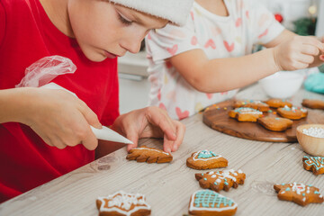 Obraz na płótnie Canvas Boy 8 years old in red Santa hat decorate Christmas gingerbread cookies with icing on wooden table in kitchen