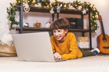 little young boy, in a yellow sweater, lies on his stomach, smiles and looks at the laptop monitor. In the background, Christmas decorations, and the twinkling of lights.