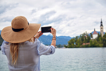 Fototapeta premium Young adult tourist woman enjoying on a lake while taking photos with a modern smartphone.