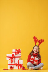cute little girl 5 years old in red deer horns, sitting with gift boxes on a yellow background in studio. child smiles happily and looks into the frame. Advertising. copy space