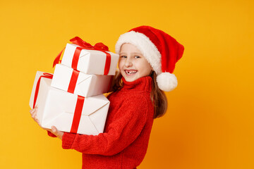 little cheerful girl in a red sweater and Santa hat holds a pyramid of Christmas gift boxes on a yellow background in the studio. child laughs and receives a lot of gifts for Christmas. Advertising.