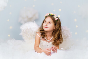 cheerful little cute girl in an angelic dress with wings and a halo lies in the clouds against the...