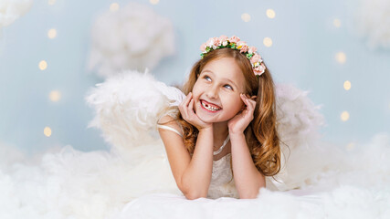 cheerful little cute girl in an angelic dress with wings and a halo lies in the clouds against the...