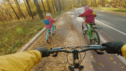 Father with her daughters have a bike ride in autumn park, POV view. Two little girls riding bicycle on bike path, family walk at beautiful sunny day, happy childhood
