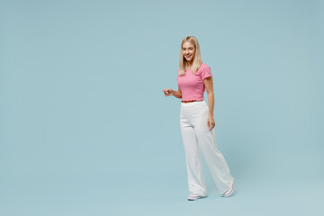 Full body side view smiling happy young blonde woman in casual pink t-shirt walking going strolling look camera isolated on plain pastel light blue background studio portrait People lifestyle concept