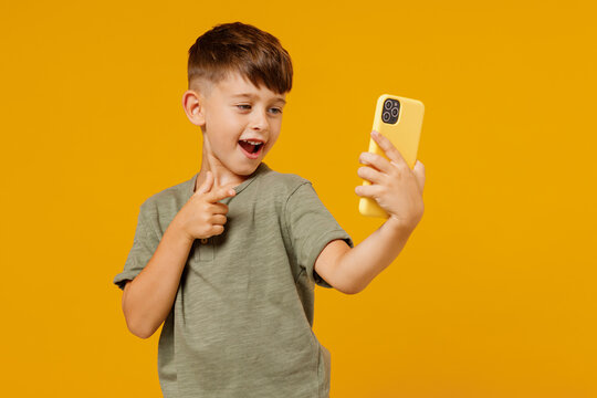 Little small smiling boy 6-7 years old in green t-shirt doing selfie shot on mobile cell phone point finger on camera isolated on plain yellow background. Mother's Day love family lifestyle concept.