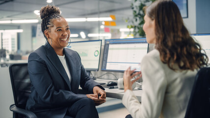 Two Female Colleagues Fondly Talk to Each Other, Laugh and Smile while Working on Computers in...