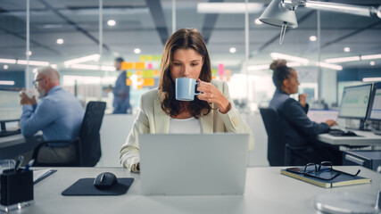 Young Happy Businesswoman Using Computer in Modern Office with Colleagues. Stylish Beautiful Manager Smiling, Working on Financial and Marketing Projects. Drinking Tea or Coffee from a Mug.